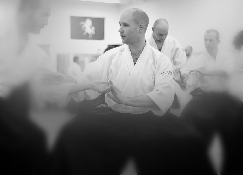 Phil Rozier seated in a keikogi, demonstrating a wrist hold while other students train behind him.